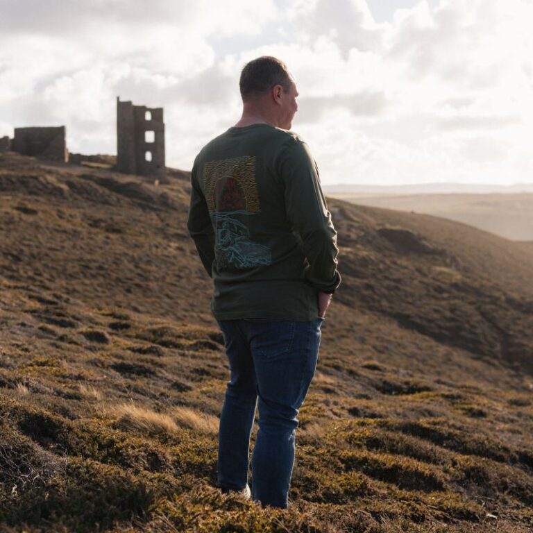 Man standing on a rugged Cornish moorland hillside looking out across the landscape at sunset, wearing a Bimbling Books & Beer long-sleeve top, with historic mine ruins in the distance, reflecting during a solo walk in Cornwall focused on slow living, mental health, and outdoor storytelling.