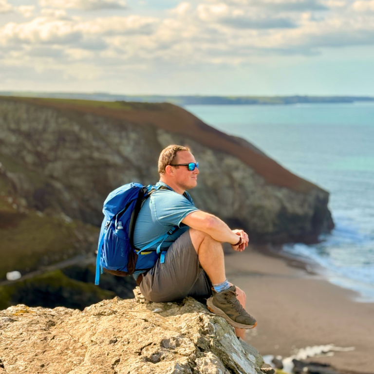 Man with a blue backpack sitting on a cliff edge overlooking a sandy beach and turquoise sea on the Cornwall coast.