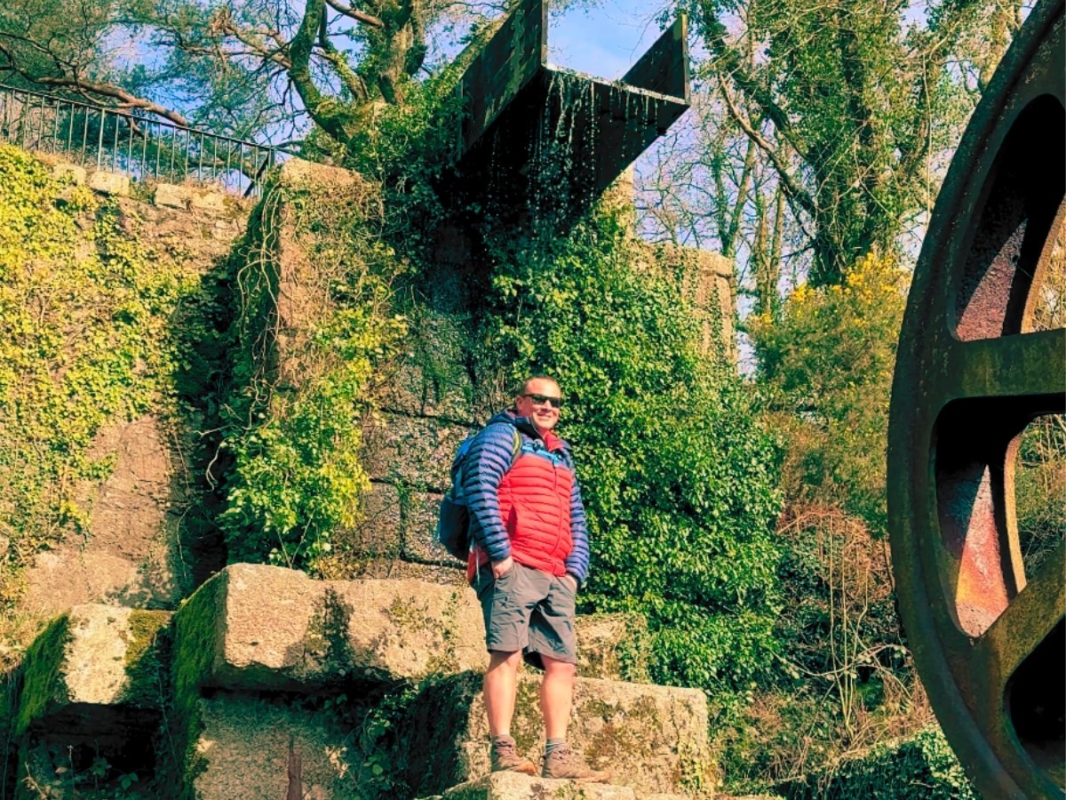Man standing in front of the water run at Carmears Wheel Pit in the Luxuylan Valley near St Austell, Cornwall. Man is dressed in a berghaus coat and oakley sunglasses smilling with his hands in pockets. hidden history