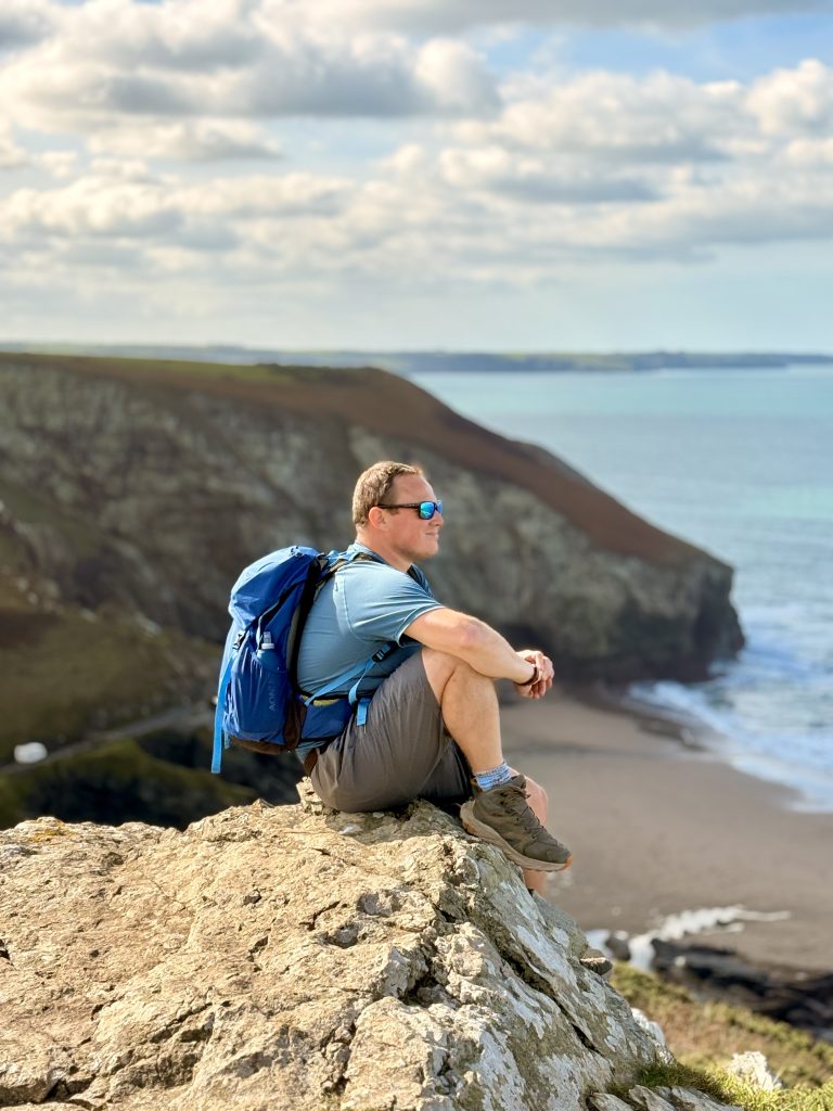 About Dan from Bimbling, Books & Beer sitting on coastal cliffs along the South West Coast Path in Cornwall during an outdoor adventure