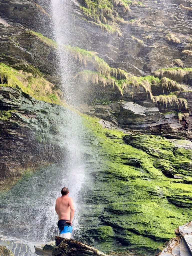 Man standing under Porth Cadjack Cove waterfall in Cornwall with water cascading down a moss covered cliff onto the beach below