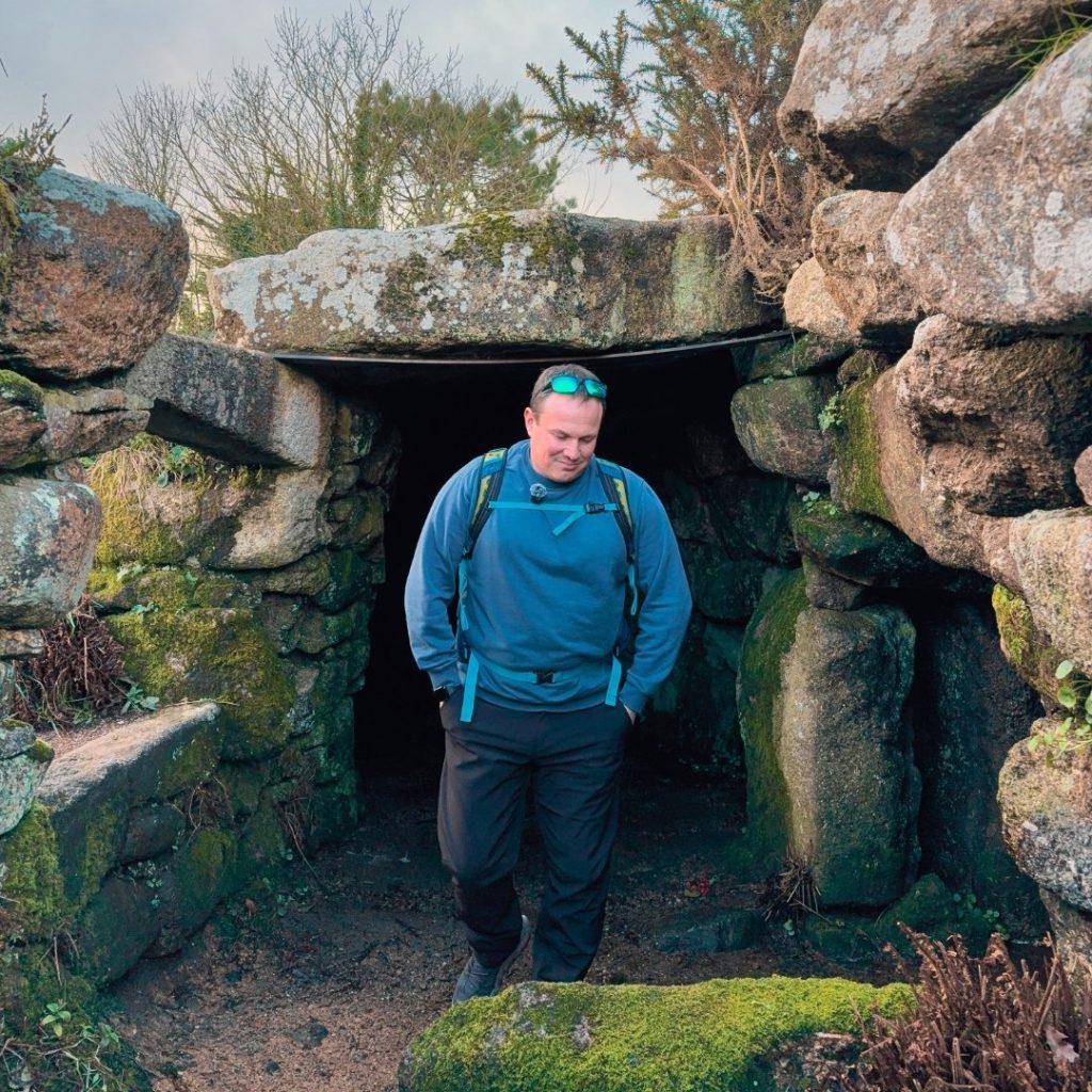 Stone entrance to an ancient settlement (Carn Euny Ancient Village) with a person emerging from a low granite structure surrounded by weathered rocks