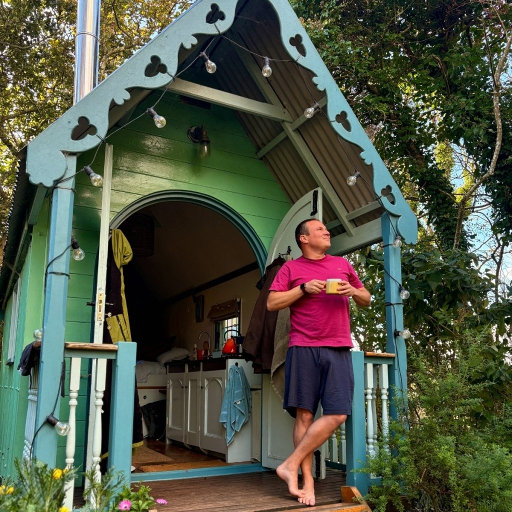 Man standing outside a cosy shepherd’s hut in woodland, enjoying a morning coffee at a peaceful countryside stay in Cornwall