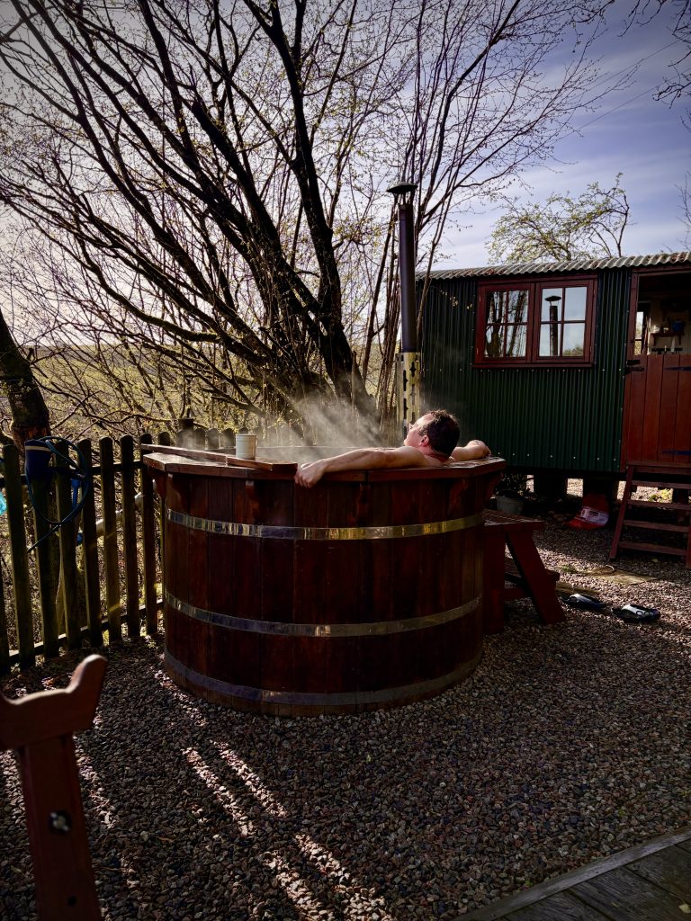 Man relaxing in a wood fired hot tub outside a shepherd’s hut stay in the Cornish countryside near Duloe, surrounded by trees and peaceful rural views