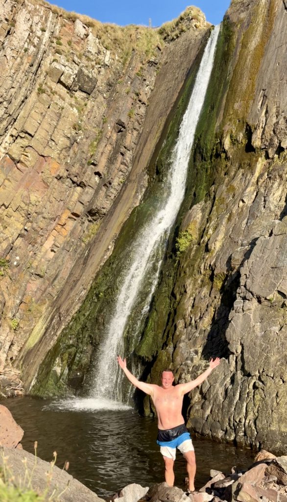 Man standing in front of Speke's Mill Mouth waterfall in North Devon with water cascading 60ft down a cliff into a plunge pool