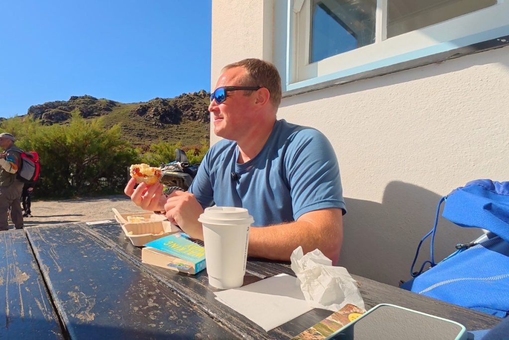 Man eating a cream tea outside Kynance Cove Cafe in Cornwall with coastal hills in the background on a sunny day.
