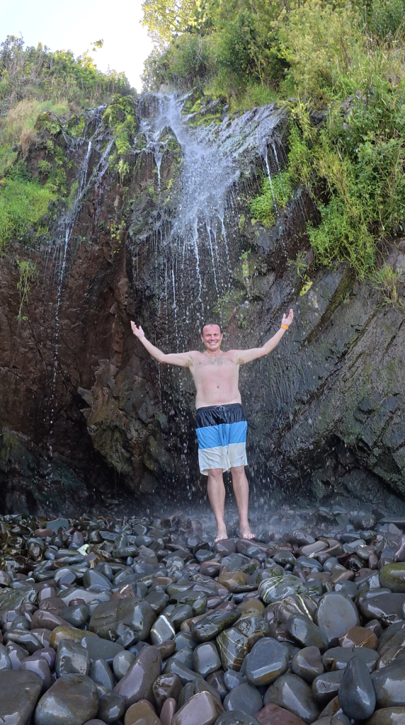 Man standing under Clovelly waterfall on a rocky beach in North Devon with water flowing over cliffs into the sea