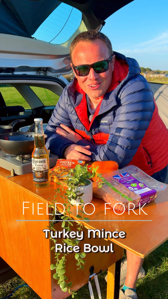 Warm sunset campsite cooking scene featuring Dan preparing a Mexican Turkey Rice Bowl from the back of a camp kitchen setup beside a TentBox, with ingredients, a Corona beer and golden evening light creating a cosy outdoor cooking atmosphere.