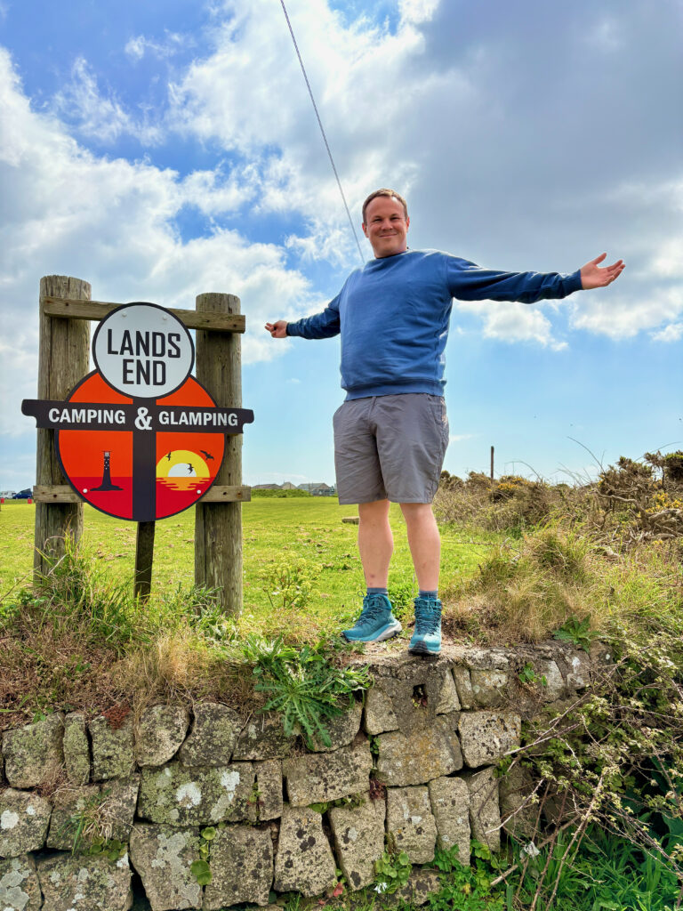 Dan standing beside the Land’s End Camping & Glamping sign in Cornwall under a dramatic blue sky, photographed during a TentBox camping trip exploring the far west coast of Cornwall.